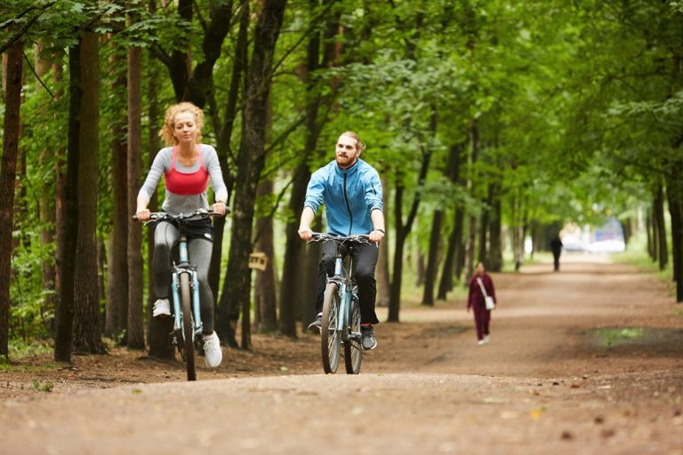 Two young active cyclists riding on bicycles along wide road between trees in park on summer day